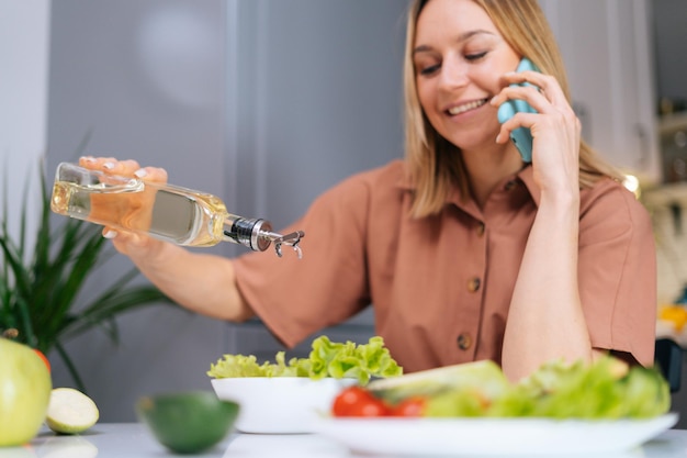 Szczęśliwa młoda kobieta nalewa oliwę z oliwek do sałatki jarzynowej i rozmawia przez telefon w kuchni z nowoczesnym wnętrzem. Pojęcie zdrowego odżywiania.