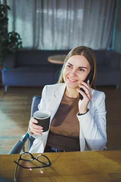 Szczęśliwa młoda kobieta o długich blond włosach w stylowych ubraniach siedzi przy stole z laptopem, popijając świeżą kawę i rozmawiając przez telefon Pracuj w nowoczesnym biurze z dużymi oknami