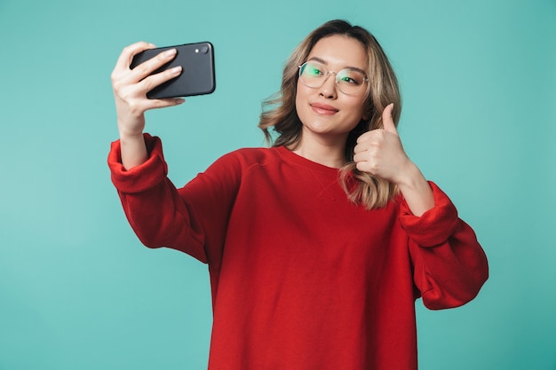 szczęśliwa młoda kobieta pozuje na białym tle nad niebieską ścianą, rozmawiając przez telefon komórkowy, zrób selfie pokazując kciuk do góry