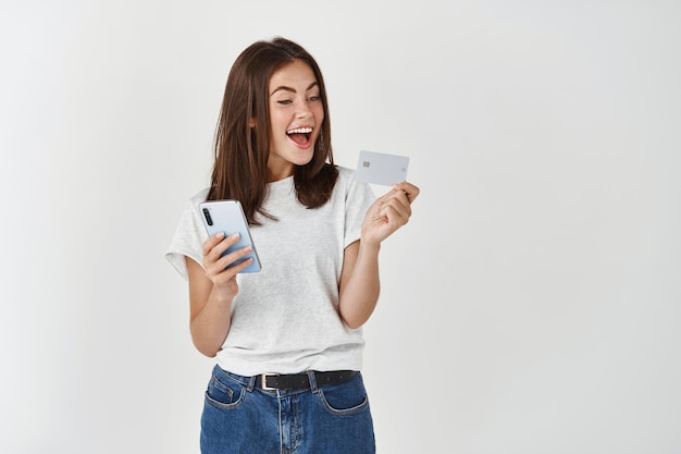 Szczęśliwa Młoda Kobieta Przy Użyciu Telefonu Komórkowego I Trzymając Plastikową Kartę Kredytową, Dokonując Zakupu, Stojąc Nad Białą ścianą.