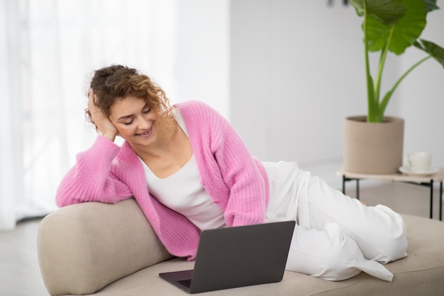 Szczęśliwa młoda kobieta relaksuje z laptopem na kanapie w domu