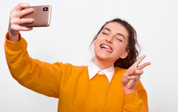 Szczęśliwa młoda kobieta robi zabawny grymas z językiem i gestem pokoju w pomarańczowym swetrze biorąc selfie na smartfonie Ładna dziewczyna studentka robi autoportret na białej ścianie Ludzie emocje