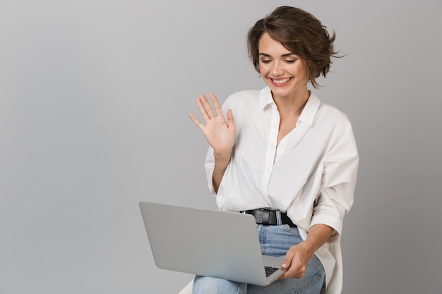 Szczęśliwa młoda kobieta siedzi na stołku na białym tle nad szarą ścianą, trzymając laptop macha