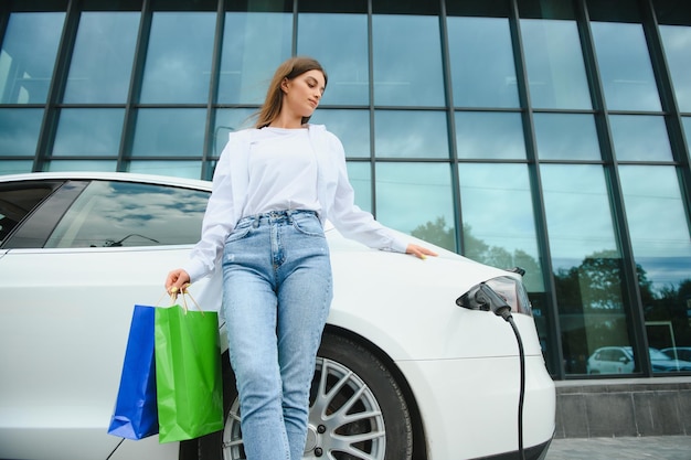 Szczęśliwa młoda kobieta stojąca na miejskim parkingu w pobliżu samochodu elektrycznego ładującego akumulator samochodowy z małej stacji miejskiej trzymającej torby na zakupy