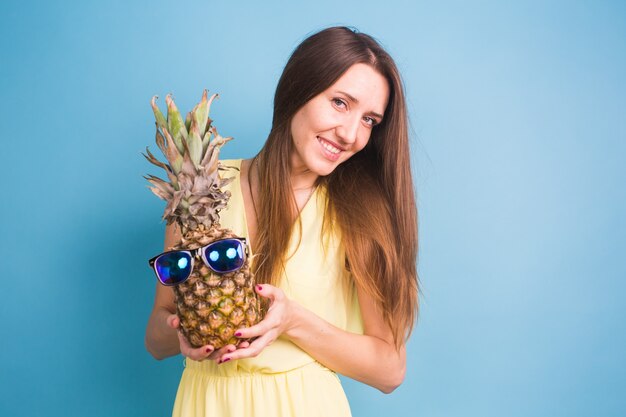 Zdjęcie szczęśliwa młoda kobieta trzyma ananasa z okularami przeciwsłonecznymi na niebieskim tle.