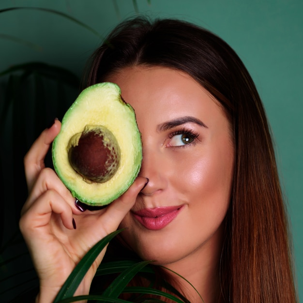 Szczęśliwa młoda kobieta trzyma dojrzałą avocado połówkę na zielonym tle.