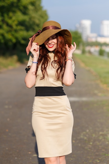 Szczęśliwa młoda kobieta trzymająca szeroki kapelusz pozuje latem w parku