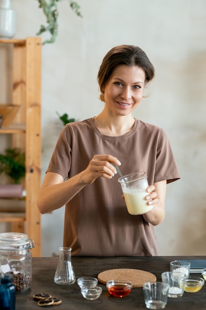 Szczęśliwa młoda kobieta trzymająca szkło z roztopioną masą mydlaną nad stołem podczas robienia mikstury do naturalnych ręcznie robionych produktów kosmetycznych