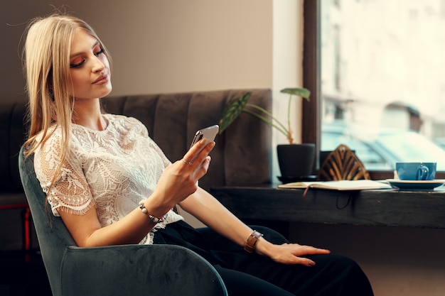 Szczęśliwa młoda kobieta używa jej smartphone podczas gdy siedzący w bufecie