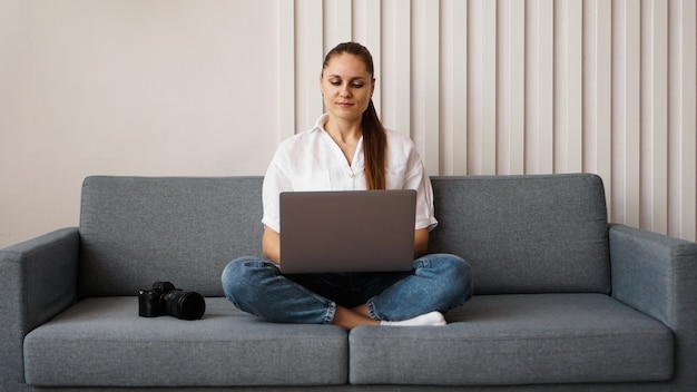 Szczęśliwa młoda kobieta używa laptopa w domu. Na kanapie obok kobiety stoi aparat. Fotograf retuszuje zdjęcia w domu.