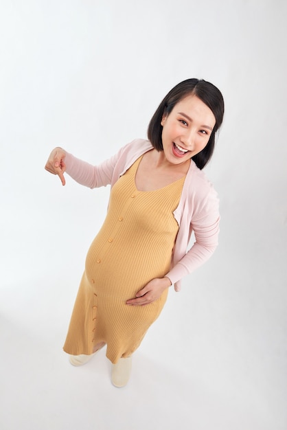 Szczęśliwa młoda kobieta w ciąży na białym tle, kobieta wskazuje palcem na brzuch