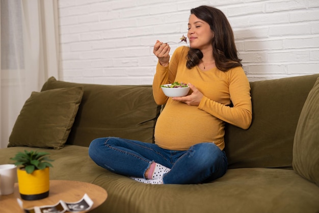 Szczęśliwa Młoda Kobieta W Ciąży Siedzi I Je Sałatkę Owocową Na Kanapie W Domu