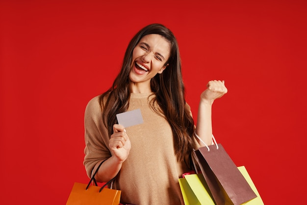 Szczęśliwa młoda kobieta w codziennej odzieży niosącej torby na zakupy i kartę kredytową