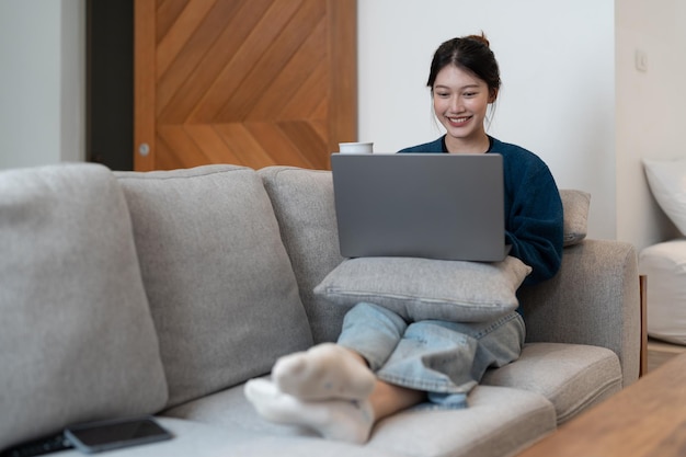 Szczęśliwa młoda kobieta w niebieskiej bluzie pracuje w domu na niebieskiej kanapie z laptopem i kotem praca zdalna i edukacja pozostając w domu