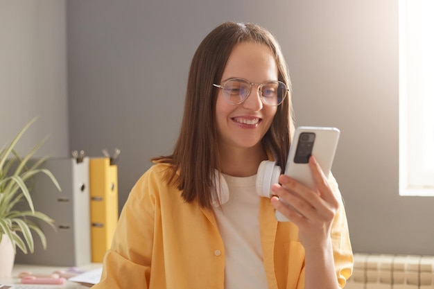 Szczęśliwa młoda kobieta w przypadkowych ubraniach z telefonem komórkowym w rękach przeglądająca smartfona siedząca w biurze w swoim miejscu pracy i sprawdzająca sieci społecznościowe podczas przerwy