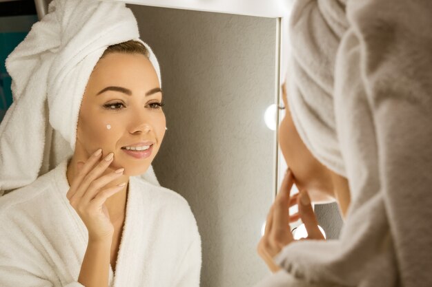 Szczęśliwa młoda kobieta w ręczniku przed lustrem nakłada krem na twarz