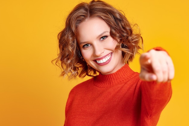 Szczęśliwa młoda kobieta w stylowym czerwonym swetrze uśmiecha się i wskazuje na kamerę na żółtym tle