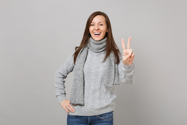 Szczęśliwa młoda kobieta w szarym swetrze, szalik pokazując znak zwycięstwa na białym tle na tle szarej ściany w studio. Zdrowy styl życia moda, szczere emocje ludzi, koncepcja zimnej pory roku. Makieta miejsca na kopię.