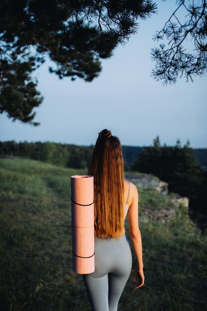 Zdjęcie szczęśliwa młoda kobieta w ubraniach fitness z matą jogową idąca na ćwiczenia w lesie o zachodzie słońca