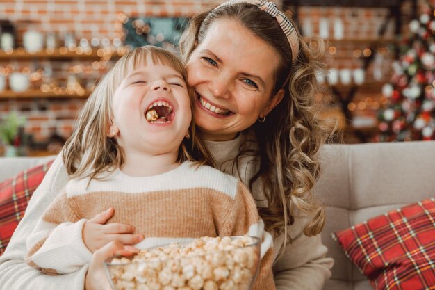 Szczęśliwa młoda mama i dziewczynka śmieją się siedząc na kanapie i jedzą popcorn. Rodzina oglądająca telewizję i jedząca popcorn.