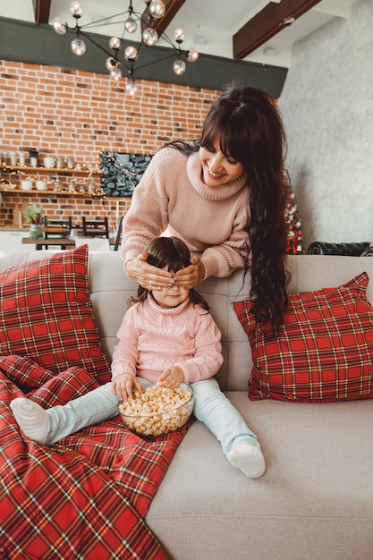 Szczęśliwa Młoda Mama I Dziewczynka śmieją Się Siedząc Na Kanapie I Jedzą Popcorn. Rodzina Oglądająca Telewizję I Jedząca Popcorn.