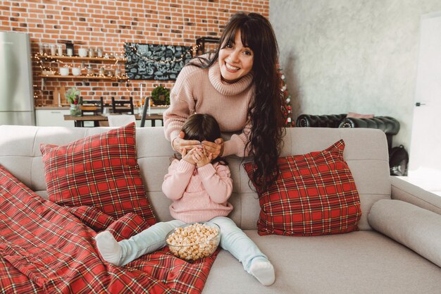 Szczęśliwa młoda mama i dziewczynka śmieją się siedząc na kanapie i jedzą popcorn. Rodzina oglądająca telewizję i jedząca popcorn.