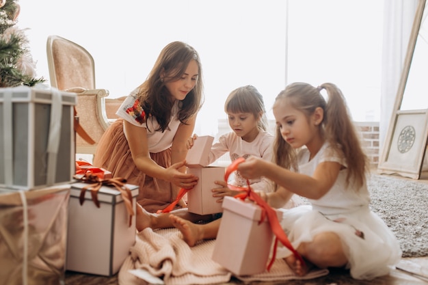 Szczęśliwa Młoda Mama I Jej Dwie Urocze Córki W ładnych Sukienkach Siedzą Na Dywanie I Otwierają Noworoczne Prezenty W Jasnym, Przytulnym Pokoju.