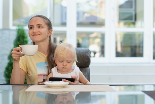 Szczęśliwa młoda mama pije kawę i trzyma dziecko w ramionach. Dziecko ogląda bajki na telefonie.