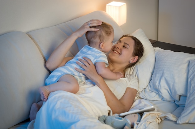 Szczęśliwa młoda matka bawi się i obejmuje swojego chłopca w łóżku późną nocą