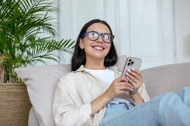 Szczęśliwa młoda nastolatka leżąca na kanapie w domu i korzystająca z telefonu odbiera wiadomości czaty z przyjaciółmi