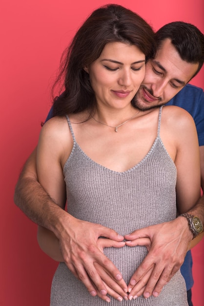 Szczęśliwa młoda para pokazująca serce z rękami na brzuchu ciężarnej żony odizolowanej na czerwonym tle