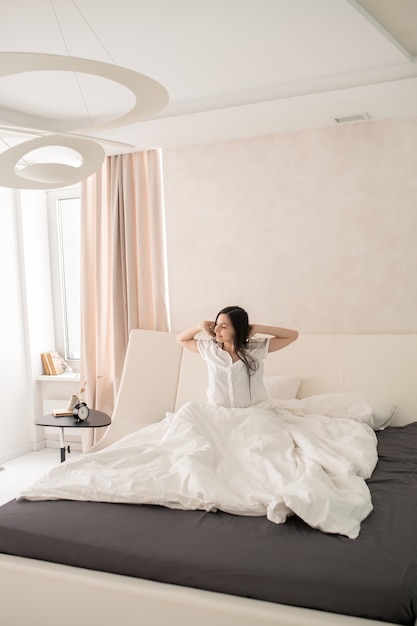 Szczęśliwa młoda piękna brunetka kobieta rozciągająca się rano na dużym wygodnym podwójnym łóżku pod białym kocem
