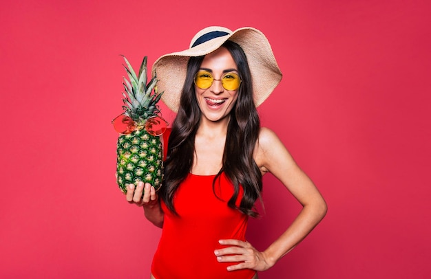 Szczęśliwa młoda piękna kobieta w okularach przeciwsłonecznych w czerwonym stroju kąpielowym i dużym letnim kapeluszu z ananasem w okularach przeciwsłonecznych w ręku pozowanie na różowym tle