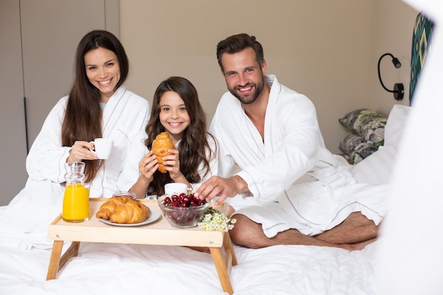 Szczęśliwa młoda piękna rodzina w szlafrokach bierze poranne śniadanie w luksusowym pokoju hotelowym