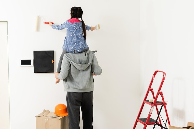 Szczęśliwa młoda rodzina remontuje swój dom, maluje ścianę wałkiem malarskim, mama stoi na drabinie i uśmiecha się.
