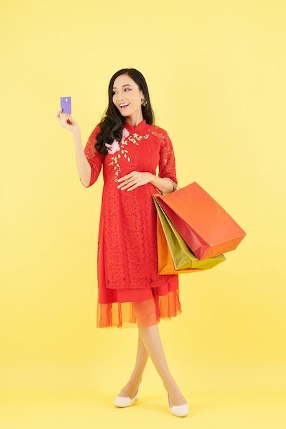 Szczęśliwa młoda Wietnamka w koronkowej czerwonej sukience, trzymająca papierowe torby i patrząca na kartę kredytową w dłoni