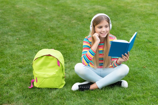 Szczęśliwa nastolatka czytająca książkę słuchająca słuchawek siedzących na trawie po odrabianiu lekcji w szkole