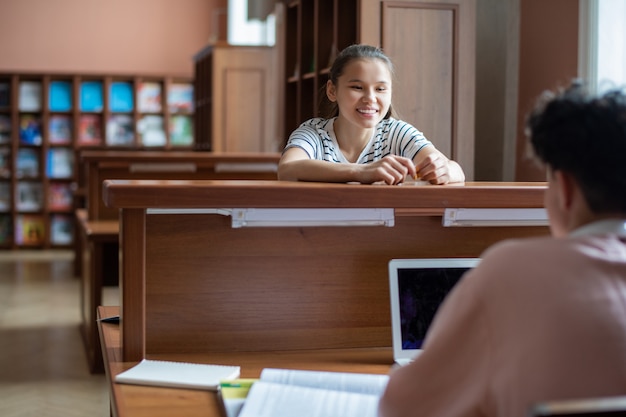 Szczęśliwa Nastolatka Patrząc Na Swojego Kolegę Z Klasy Z Laptopem Podczas Rozmowy W Bibliotece Uczelni Po Zajęciach