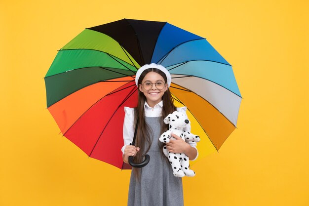 Szczęśliwa nastolatka w okularach i berecie pod kolorowym parasolem dla ochrony przed deszczem w sezonie jesiennym trzymaj zabawkę z dzieciństwa