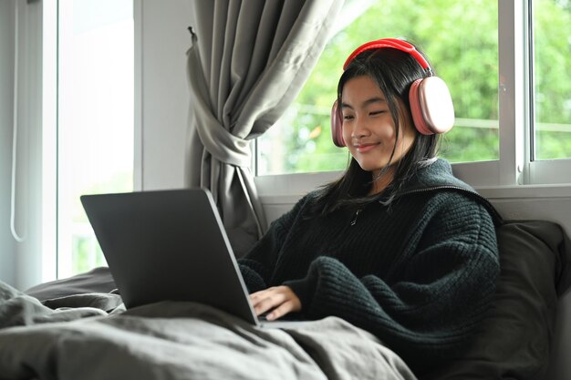 Szczęśliwa nastolatka w słuchawkach oglądająca film lub surfująca po internecie na laptopie siedząc w sypialni