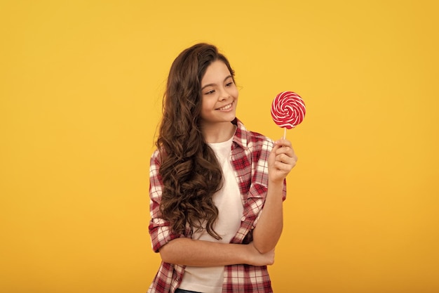 Szczęśliwa nastolatka z długimi kręconymi włosami trzyma lizak karmelowy cukierek na żółtym tle sklep ze słodyczami