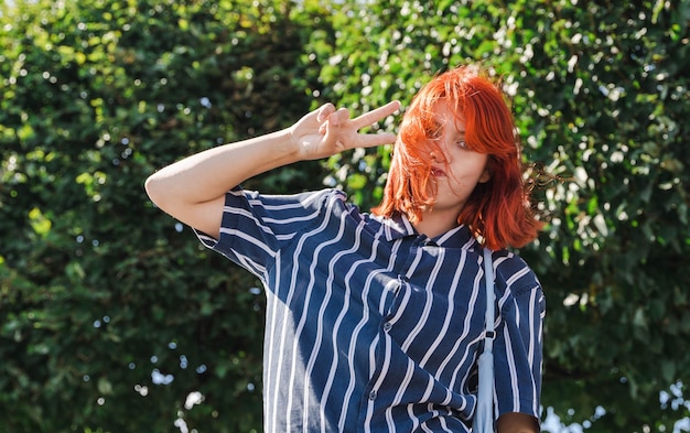 Szczęśliwa nastolatka z pokolenia Z z czerwonymi włosami pokazuje znak zwycięstwa w parku miejskim
