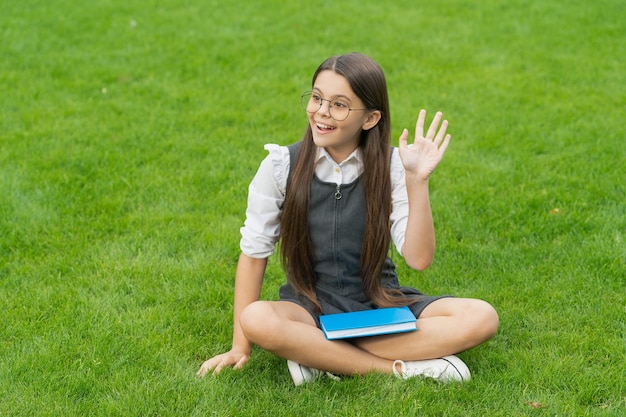 Szczęśliwa nastoletnia uczennica macha ręką siedzącą na trawie Cześć szkoła Powrót do szkoły