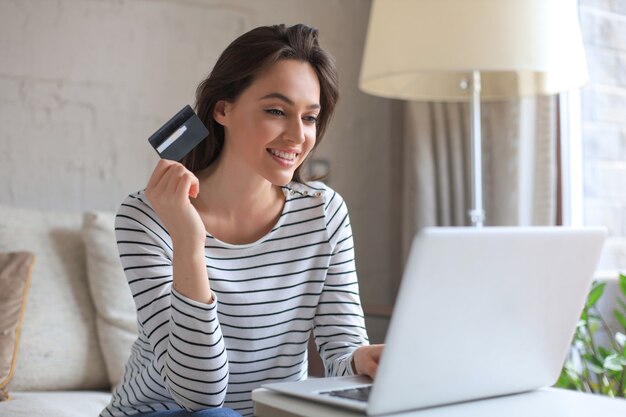 Szczęśliwa naturalna brunetka za pomocą karty kredytowej i laptopa w salonie.