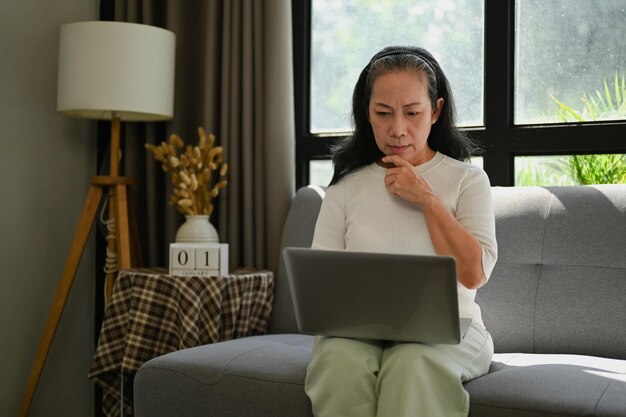 Szczęśliwa pani w średnim wieku sprawdza pocztę e-mail lub pracuje na laptopie, relaksując się na kanapie w domu