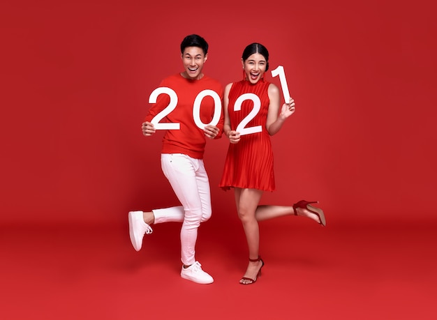 Szczęśliwa para azjatyckich w czerwonym stroju casual pokazująca numer 2021 powitanie szczęśliwego nowego roku z uśmiechem na jasnoczerwonym.