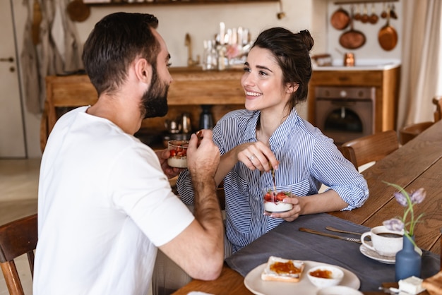 szczęśliwa para brunetka mężczyzna i kobieta razem jedzą deser panna cotta siedząc przy stole w domu