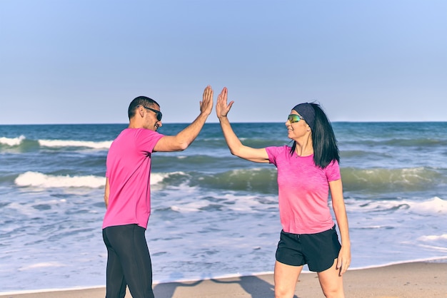 Szczęśliwa para fit świętuje po zakończeniu treningu na plaży. Różowe koszule i czarne spodenki. Morze w tle.