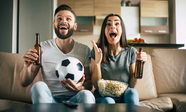 Szczęśliwa para kibiców ogląda mecz piłki nożnej w telewizji z przekąskami, piwem i piłką na kanapie