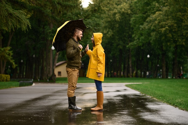 Szczęśliwa para miłości przysięga sobie nawzajem w parku, letni deszczowy dzień. Mężczyzna i kobieta pod parasolem w deszczu, romantyczna randka na ścieżce spacerowej, deszczowa pogoda w alei